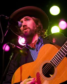 a man with a hat playing an acoustic guitar in front of microphones and lights