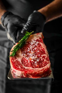 a person in black gloves holding up a piece of meat with herbs on top and salt on the side