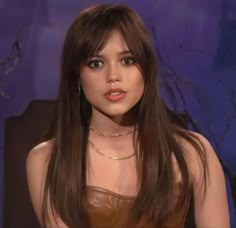 a woman with long brown hair sitting in front of a blue wall and wearing a leather dress