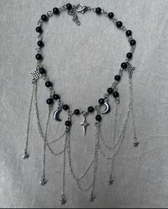 The necklace has a celestial-inspired design, featuring black beads connected by silver chain links. It includes various hanging silver charms, such as crescent moons, stars, and a central starburst or compass-shaped charm. Several delicate silver chains drape down in layers, adding an elegant, flowing effect to the piece. The overall look is both mystical and stylish, blending dark tones with metallic details for a balanced, striking appearance. Chinese Goddess, Jewelry Goth, Goth Necklace, Beaded Jewellery, Silver Chains, Chain Links, Dream Jewelry, Black Beads, Silver Charms