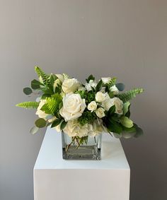 a vase filled with white flowers and greenery on top of a small table in front of a gray wall