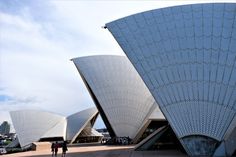 two people are walking in front of the architecture of sydney opera house, which is designed to look like it's made out of metal