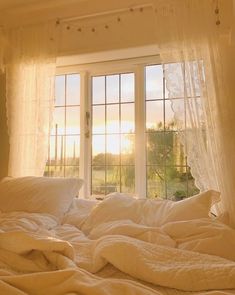 an unmade bed in front of a window with sheer curtains on the windowsill