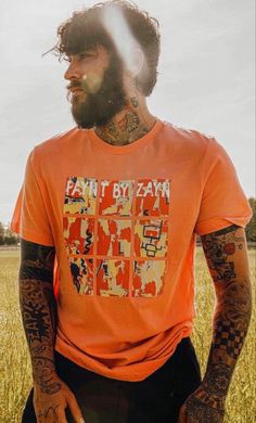 a man with long hair and tattoos standing in a field wearing an orange t - shirt