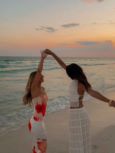 two beautiful women standing on top of a sandy beach next to the ocean at sunset