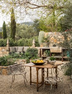 an outdoor table with fruit on it in the middle of a gravel area surrounded by trees and bushes