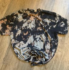 an old black and white shirt sitting on top of a wooden floor with paint splatters all over it