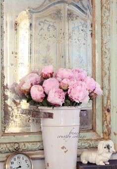 pink flowers are in a white bucket next to a small dog and an antique mirror