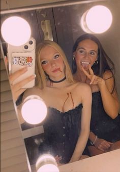 two women taking selfies in front of a mirror with light bulbs on the side