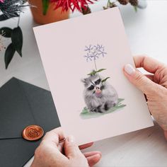 a person holding up a card with a cat on it and flowers in the background