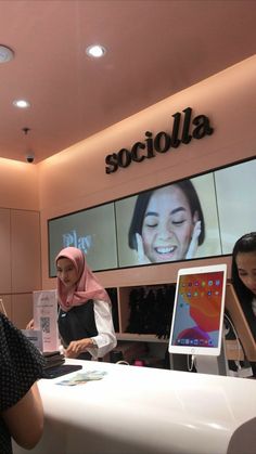 two women standing at a counter in front of a screen with a woman's face on it