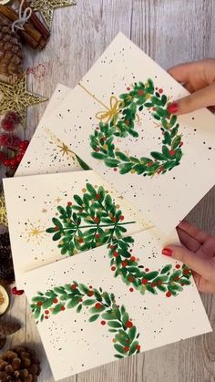 three christmas cards with holly wreaths on them and pine cones in the background, one being held up by two hands