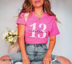 a woman sitting on the floor with flowers in her hand and wearing a pink shirt that says 13 hale