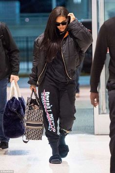 a woman walking through an airport carrying a handbag and wearing all black with her name written on the side