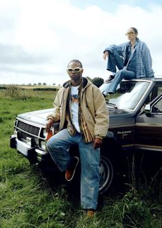 two men sitting on the hood of a car in a grassy field with one man standing next to it