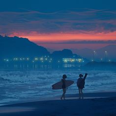 two people walking on the beach with their surfboards at sunset or dawn, one holding a surfboard