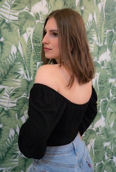 a woman with long hair wearing a black off the shoulder top and denim skirt standing in front of a wallpapered background