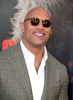 a bald man wearing sunglasses and a suit jacket smiling at the camera while standing in front of a movie poster