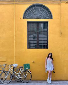 a woman standing in front of a yellow building with two bikes parked next to it