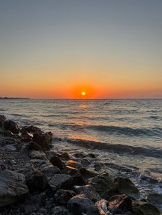 the sun is setting over the ocean with rocks on the shore and water in the foreground