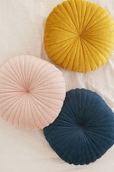 three circular pillows on a bed with white sheets and blue, yellow and pink colors