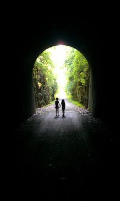 two people standing in the middle of a tunnel with trees on both sides and light at the end