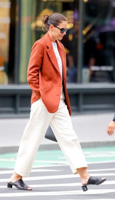 a woman walking across a cross walk wearing an orange blazer and white pants with black mules