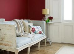 a living room with a white couch and red wall in the background, next to a table with flowers on it