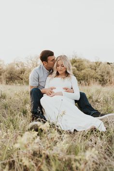 a man and woman sitting in the grass with their arms around each other while they are cuddling