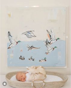 a baby is laying in a basket with ducks on the wall behind it and an art piece hanging above