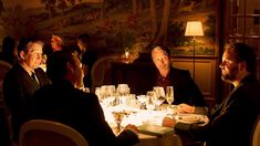 three men sitting at a dinner table with wine glasses on the table and one man standing in front of them