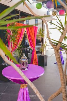 a purple table with a lantern on it in front of pink drapes and potted plants