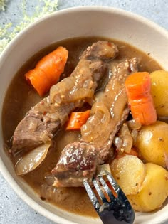 a white bowl filled with stew and carrots next to a fork on top of a table