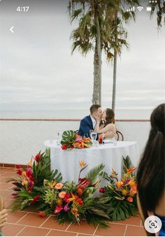 a man and woman sitting at a table with flowers on it in front of the ocean