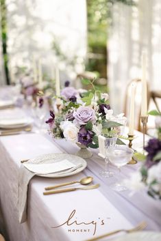 the table is set with white plates and silverware, purple flowers, greenery and candles