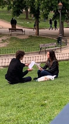 two people sitting on the grass reading books in a park with trees and benches behind them