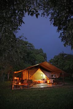 a tent is lit up at night in the grass with chairs and tables under it