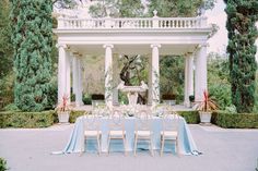 an outdoor dining table set up with blue linens and white chairs in front of a gazebo