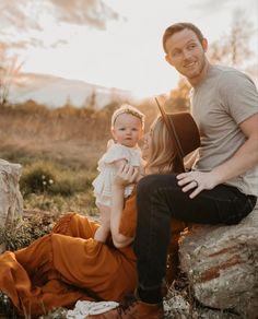 a man holding a baby while sitting on top of a rock in front of a woman