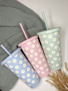 three different colored cups with straws and flowers on them next to a gray blanket