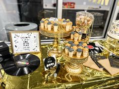 a table topped with lots of cakes and cupcakes on top of gold foil