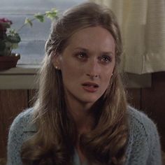 a woman with long hair wearing a sweater and looking at the camera while sitting in front of a window