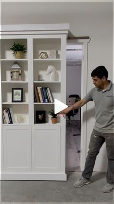 a man standing in front of a book shelf holding a white object with one hand