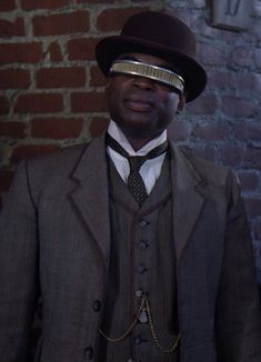 a man wearing a suit and tie with a hat on his head, standing in front of a brick wall