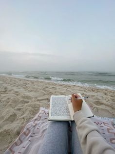 a person sitting on the beach with a book and pen in their lap looking at the ocean