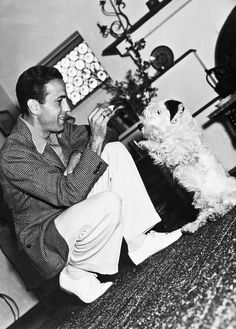 a black and white photo of a man sitting on the floor playing with his dog