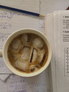 a cup of tea with ice cubes in it next to an open book and pencil