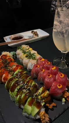 sushi is arranged on a black plate next to a glass of water and a drink