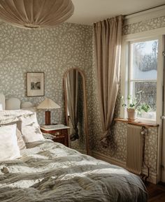 a bed sitting under a window next to a mirror and lamp in a room with floral wallpaper