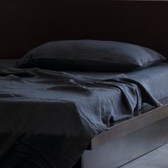 an unmade bed with black linens in a dark colored room, next to a window